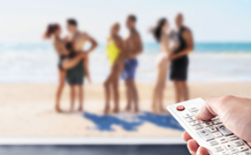 A hand holds a remote pointed at a television with a reality dating show on the screen.