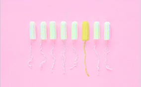 Eight tampons outside of their applicator sit in a row against a pink background and one is yellow.