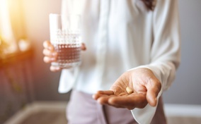 A person holds a glass of water in their right hand, and two pills in their left hand, which is in the foreground.