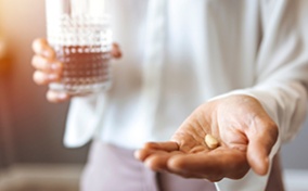 A person holds a glass of water in their right hand, and two pills in their left hand, which is in the foreground.