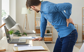 A man holds onto his lower back in pain while leaning on his computer desk.