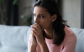 A concerned woman sits on a sofa with her hands held together against her chin.