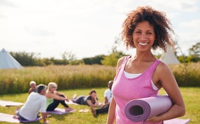 A woman smiles at the camera, wearing a pink tank top and holding an exercise mat.