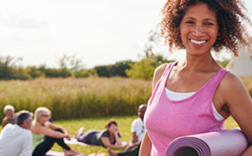 A woman smiles at the camera, wearing a pink tank top and holding an exercise mat.