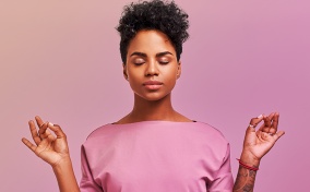 A woman in a pink shirt meditates with her eyes closed against a pink background.