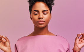 A woman in a pink shirt meditates with her eyes closed against a pink background.