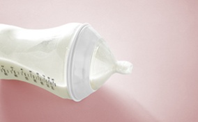 A baby bottle with milk lays on its side against a pink background.