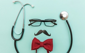 A pair of glasses and a mustache lay against a mint background with a bowtie beneath and surrounded by a stethoscope.