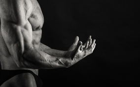 A muscled man flexes his arms out in front of him against a black background.