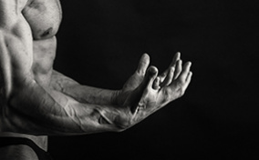A muscled man flexes his arms out in front of him against a black background.