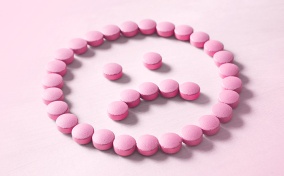 A frowning face made out of pink pills lays against a lighter pink surface.