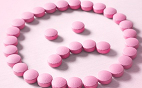 A frowning face made out of pink pills lays against a lighter pink surface.