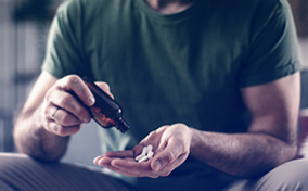 A man pours white pills from a brown bottle into his hand while sitting down.