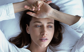 A woman lies in bed with her arms against her head.