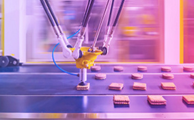 A manufacturing machine grabs a processed cookie sandwich from a conveyer belt.