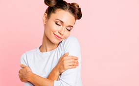 A person wearing their hair in buns closes their eyes and hugs themselves in front of a pink background.