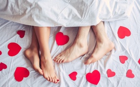 Two pairs of feet peek out from under white sheets surrounded by red hearts. 