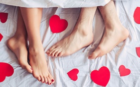 Two pairs of feet peek out from under white sheets surrounded by red hearts. 