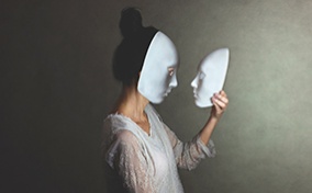 A woman in a white mask holds another mask in her hand and looks at it face to face.