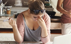 A woman holding a pen in one hand tries to concentrate and rests her forehead in her other hand, a laptop, calculator, and paper on the table in front of her. 