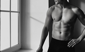 A muscled man in boxer briefs stands shirtless next to a window.