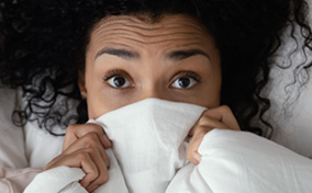 A woman lies in bed with a blanket over the lower part of her face with her eyes wide in fear.