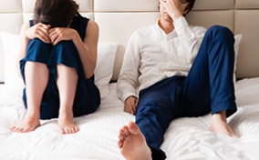 A man and woman sit clothed in bed holding their faces.