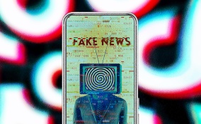 A cellphone has a person with a TV in place of their head and the words FAKE NEWS showing on screen.
