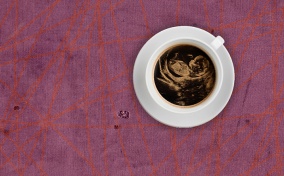A white mug of coffee sits on a purple and red lined table and shows a fetal ultrasound in the foam. 