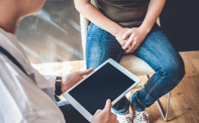 A man sits while a doctor stands in front of him holding a tablet.