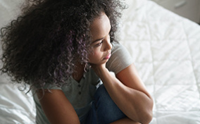 A woman sits on her bed with her leg curled up, holding her head with her hand.