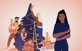 A woman stands away from her family surrounding the Christmas tree with her arms wrapped around her chest.