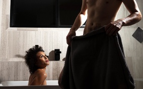 A woman sits in a bath looking up at a man standing near in only a towel.