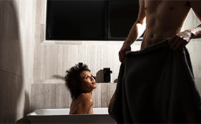 A woman sits in a bath looking up at a man standing near in only a towel.