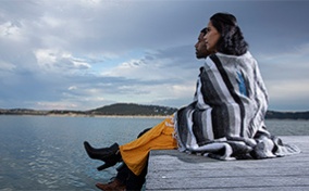 A married couple sits on the edge of a wooden dock looking out over a lake with a blanket around them.