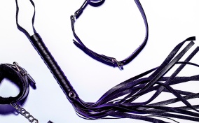 A leather whip lays between a pair of bondage cuffs and a ball gag.