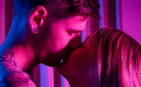 A man and woman kiss as a pink light shines on them.