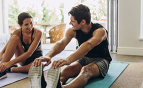 Two people sit on exercise mats stretching and smiling at each other.