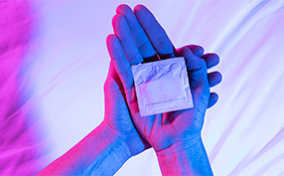 One hand holds the back of another with a wrapped condom in its palm.