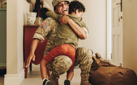 A uniformed army mom smiles as she crouches down to hug her child.