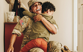 A uniformed army mom smiles as she crouches down to hug her child.