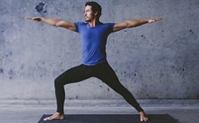 A man in black pants and a blue shirt does the Warrior II yoga pose on a black mat.
