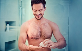 A shirtless man smiles in his bathroom while he pours a pill into his hand from a bottle.