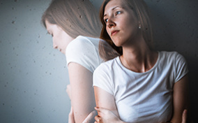 A woman leans against a wall looking to her right as a translucent image of herself turns in the same direction.