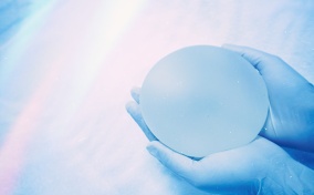 Two gloved hands hold a breast implant in their palms.