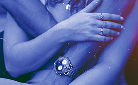 A woman holds onto the arms of a man which are wrapping around her from behind.