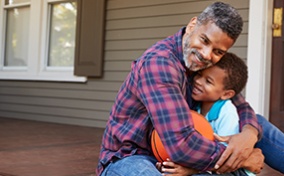 A dad sits on a front porch and hugs his child, who is holding a basketball. 
