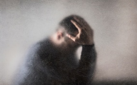A man on the other side of a fogged glass, holds his head in his hands.