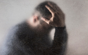 A man on the other side of a fogged glass, holds his head in his hands.