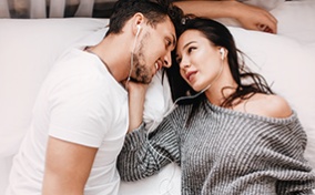 A couple lays in bed looking at each other listening to music on the same pair of earbuds.